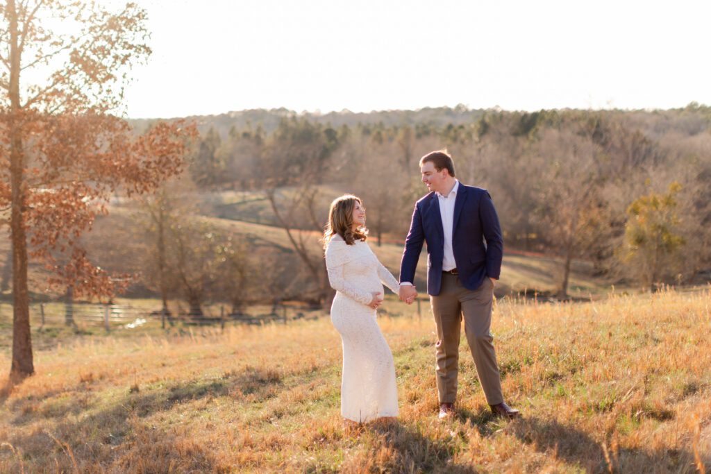 Expectant parents holding hands and smiling at each other during a golden-hour maternity session in a scenic Alabama field at SpringHouse.