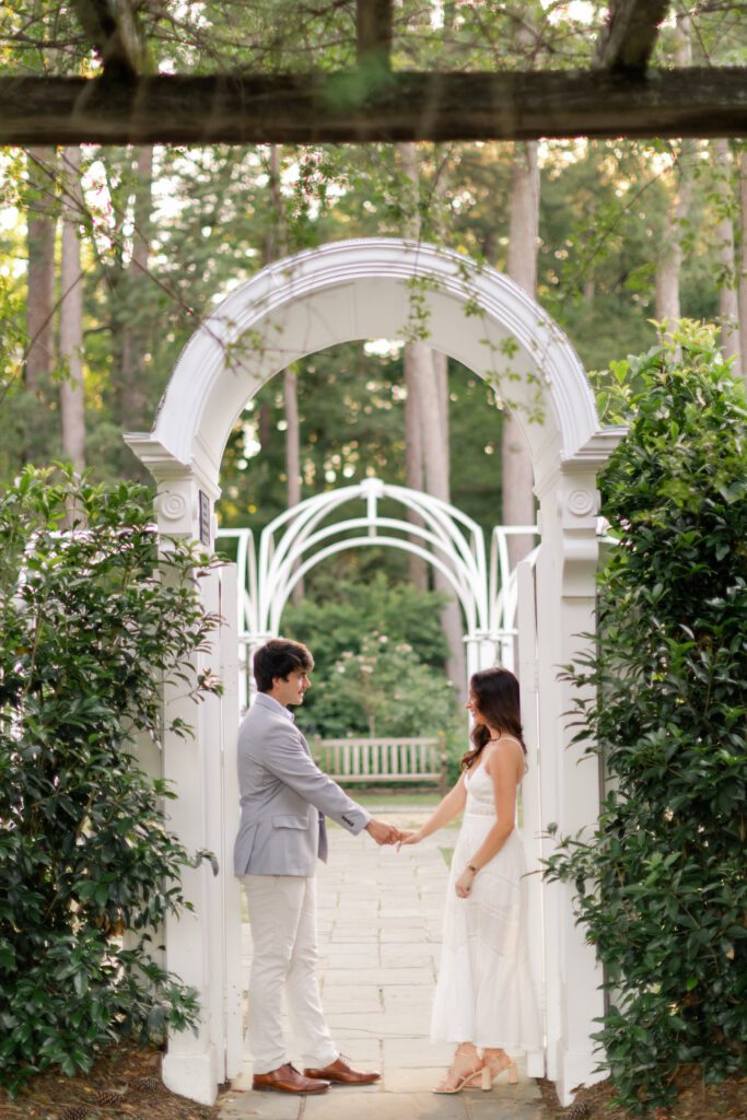 Emily and Brody hold hands beneath a white garden arch at Birmingham Botanical Gardens.