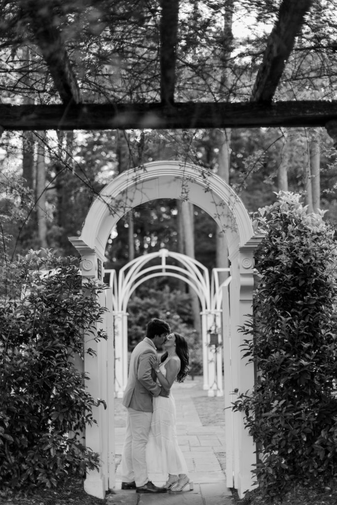 Black-and-white portrait of Emily and Brody kissing beneath a white garden arch.