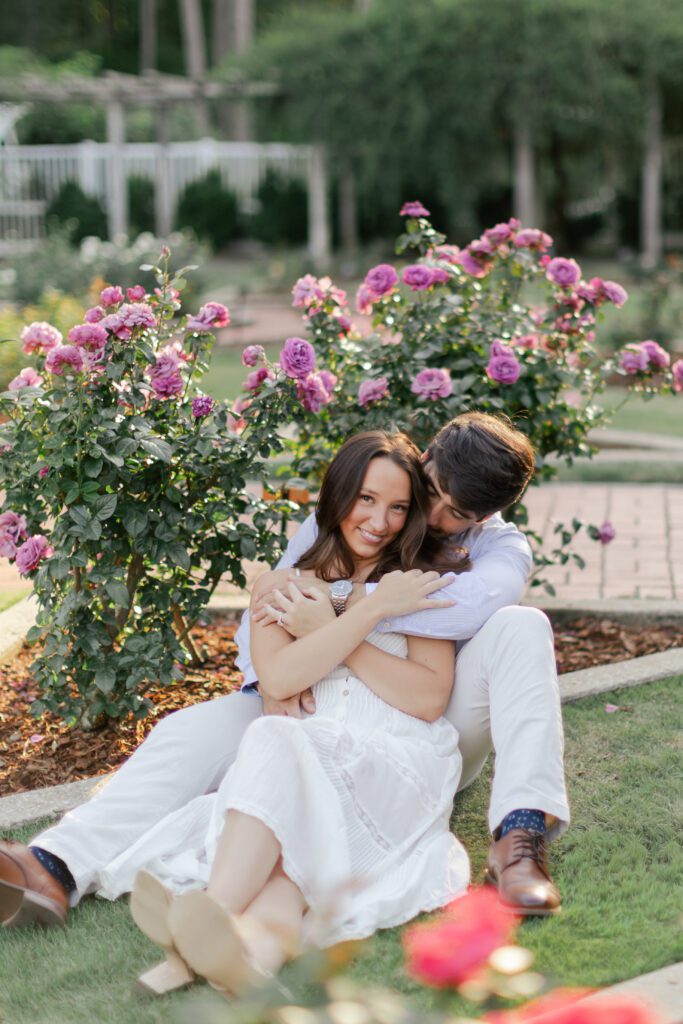 Emily and Brody sit together on the grass beside blooming purple roses.
