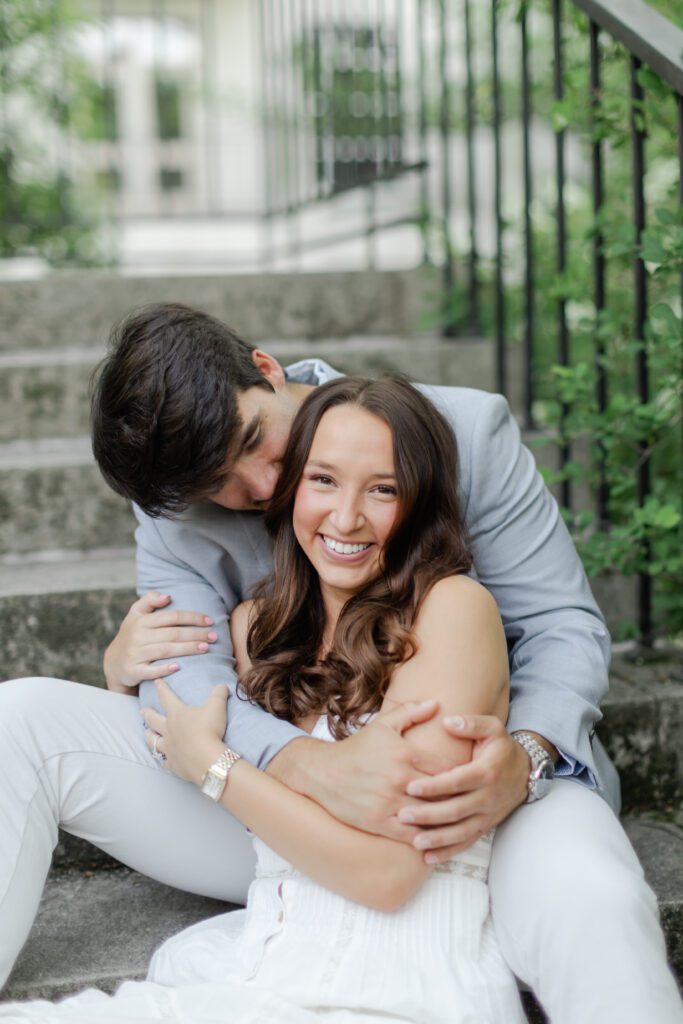 Emily smiles at the camera as Brody embraces her while they sit together on garden steps.