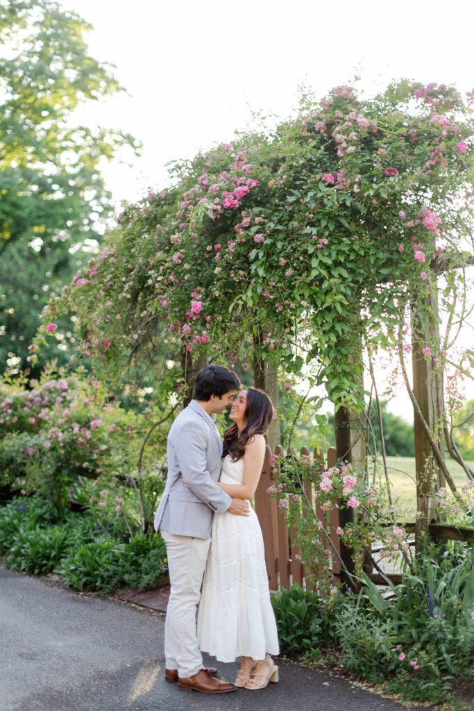 Couple embracing beneath an arch of blooming pink roses in a lush garden.