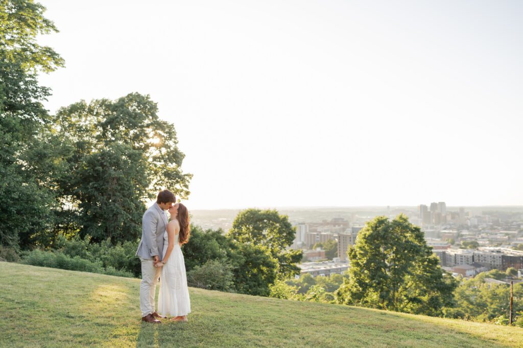 Couple kissing on a grassy overlook with a city skyline in the background at sunset.