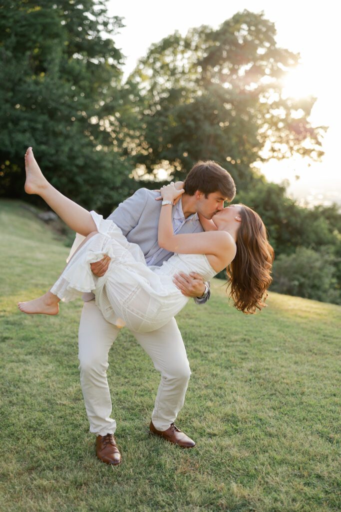 Couple kissing as one partner holds the other in his arms on a sunlit grassy hill.