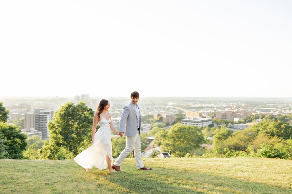 Couple walking hand in hand across a grassy overlook with the city skyline behind them.