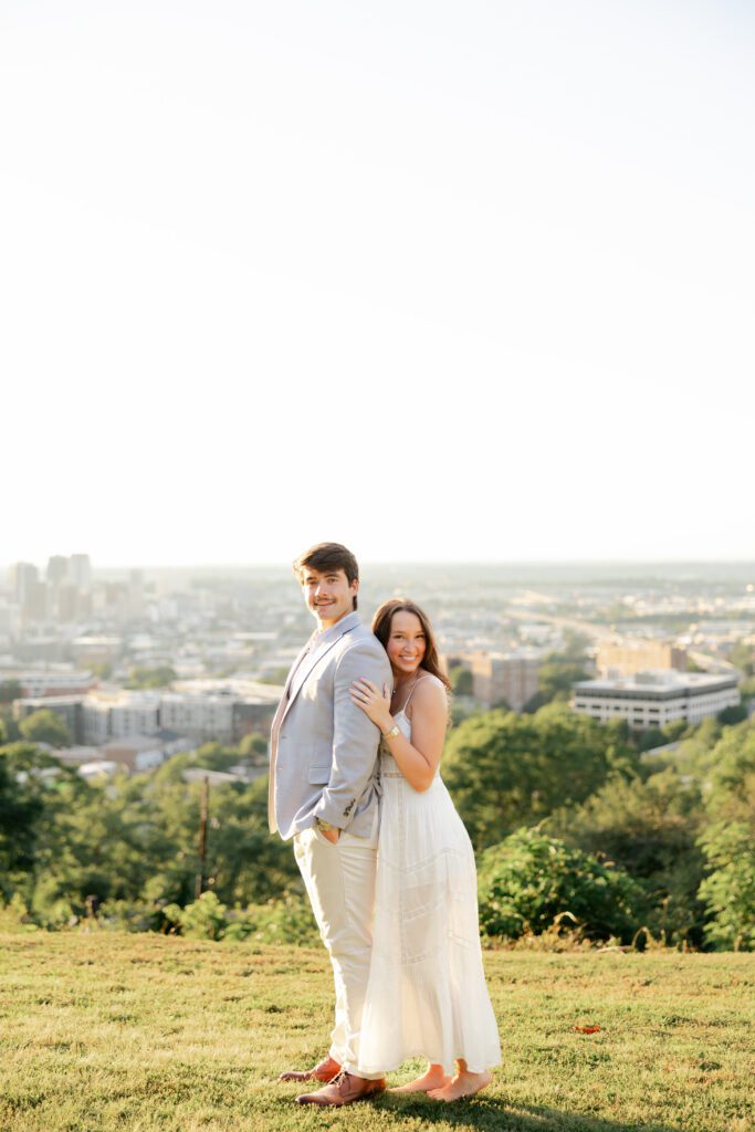 Couple posing together on a grassy hill with a softly blurred city skyline in the background.