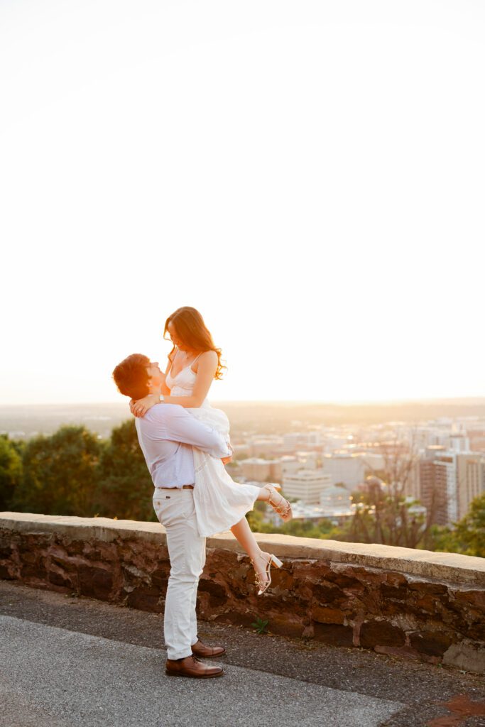 Couple embracing as one partner lifts the other beside a stone overlook at sunset.
