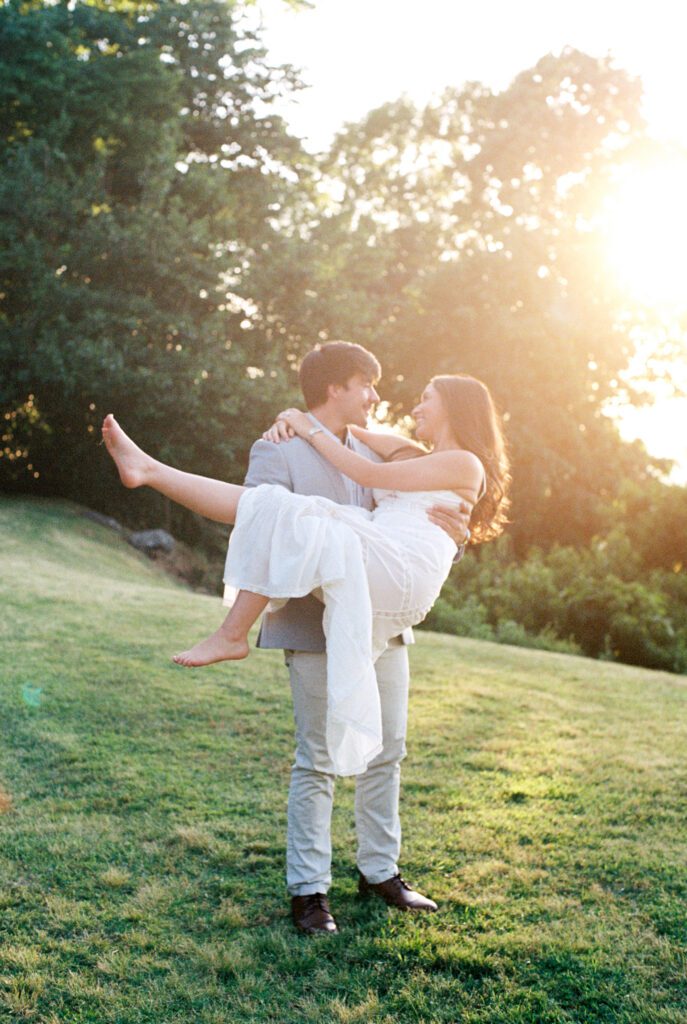 Couple laughing together while one partner carries the other across a grassy hill at golden hour.