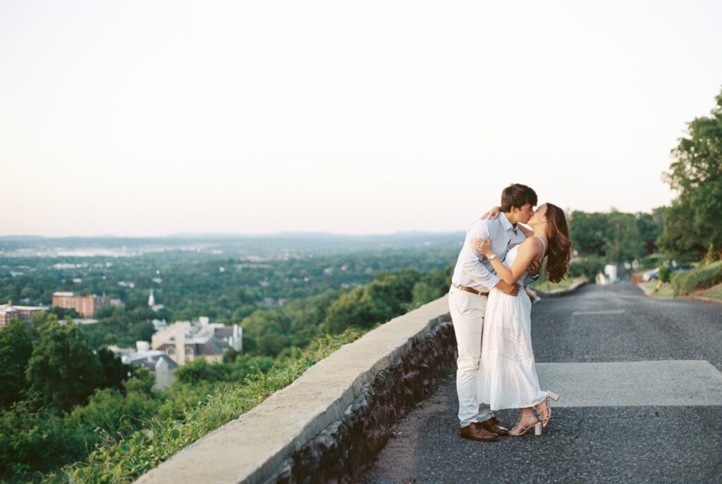 Couple kissing beside a stone overlook with a sweeping landscape in the background.