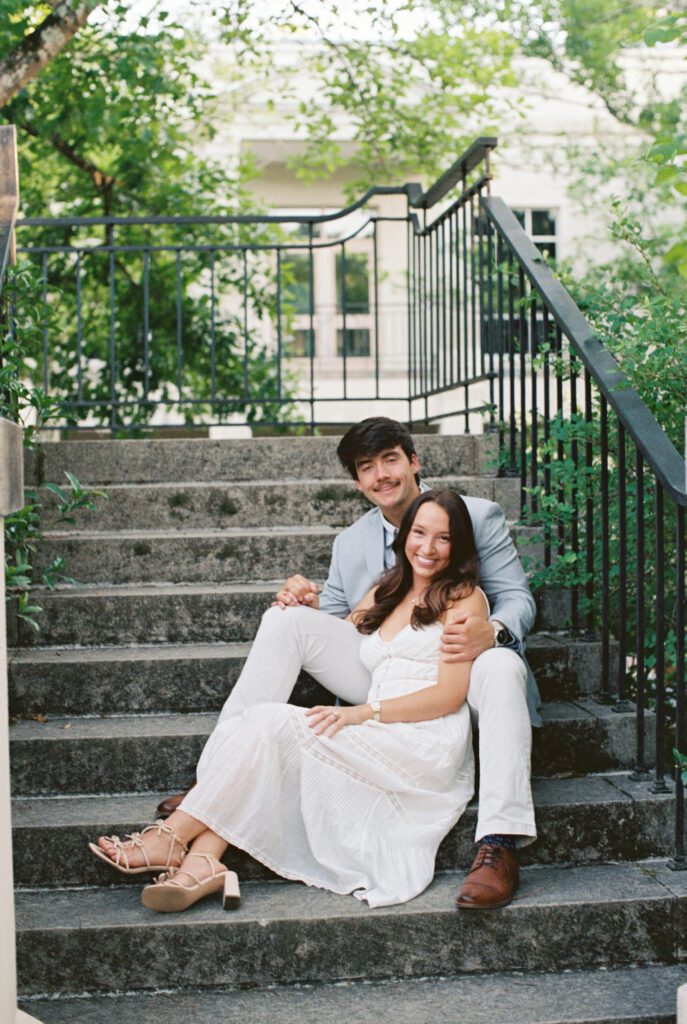 Couple sitting together on outdoor stone steps surrounded by leafy trees and black railings.