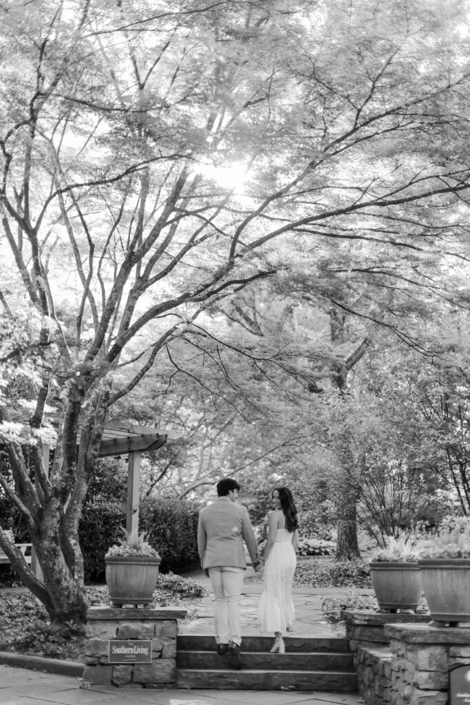 Emily and Brody walk hand in hand beneath mature trees at Birmingham Botanical Gardens.