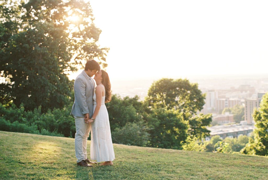 Couple standing close and holding hands on a grassy overlook with a city skyline at golden hour.