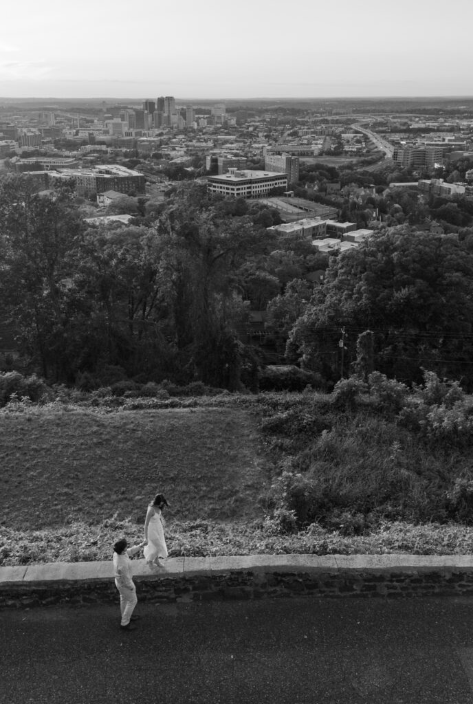 Black-and-white view of a couple walking beside a stone wall overlooking a sprawling city skyline.