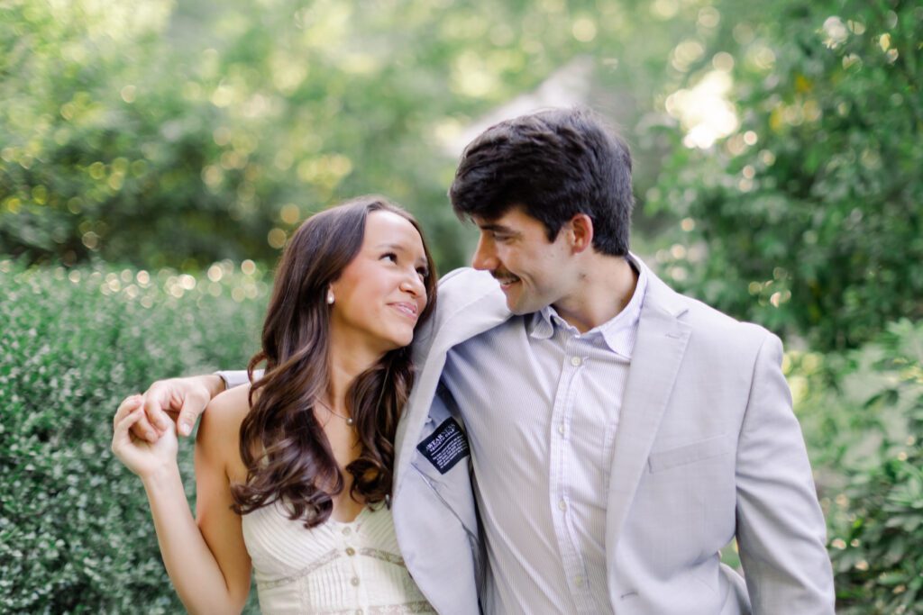Emily and Brody smile at each other surrounded by lush greenery during their Birmingham engagement photos.